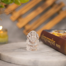 Load image into Gallery viewer, Clear Quartz Ganesh crystal figurine placed on round marble surface next to a sacred book, blurred background with candle