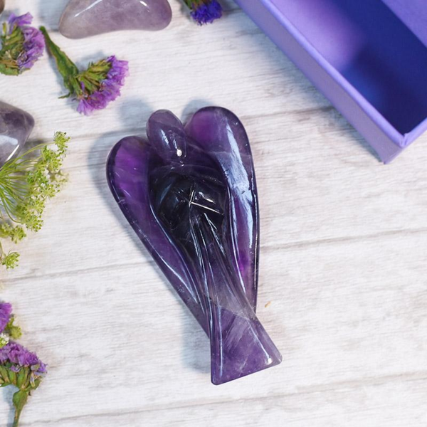 Amethyst Angel crystal figurine on a white table with soft-focus amethyst stones behind it.