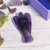Amethyst Angel crystal on a clean white table with scattered amethyst stones in the background.
