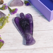 Load image into Gallery viewer, Amethyst Angel crystal on a clean white table with scattered amethyst stones in the background.
