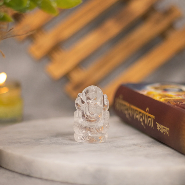 Clear Quartz Ganesh crystal figurine placed on round marble surface next to a sacred book, blurred background with candle 