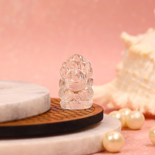 Clear quartz Ganesh figurine displayed on round marble and wooden platforms with pearls and a shell in soft pink background