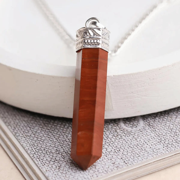 Close-up of a Red Jasper pencil pendant with a silver cap displayed against a white round stand and textured surface.