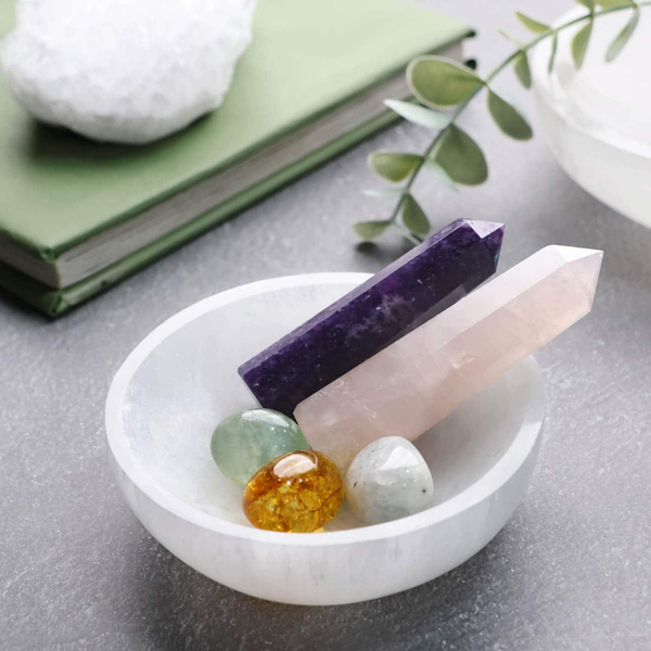 Selenite charging bowl holding amethyst, rose quartz, citrine, and other healing crystals on a grey surface