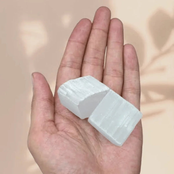 Selenite raw crystal cluster resting on the palm against a soft pink background, symbolizing cleansing and clarity.